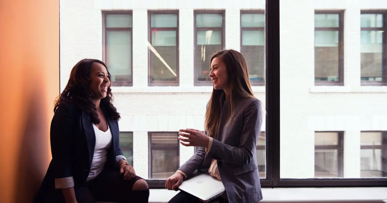 Two women sit by a large window in an office, smiling and talking while one gestures during the conversation.