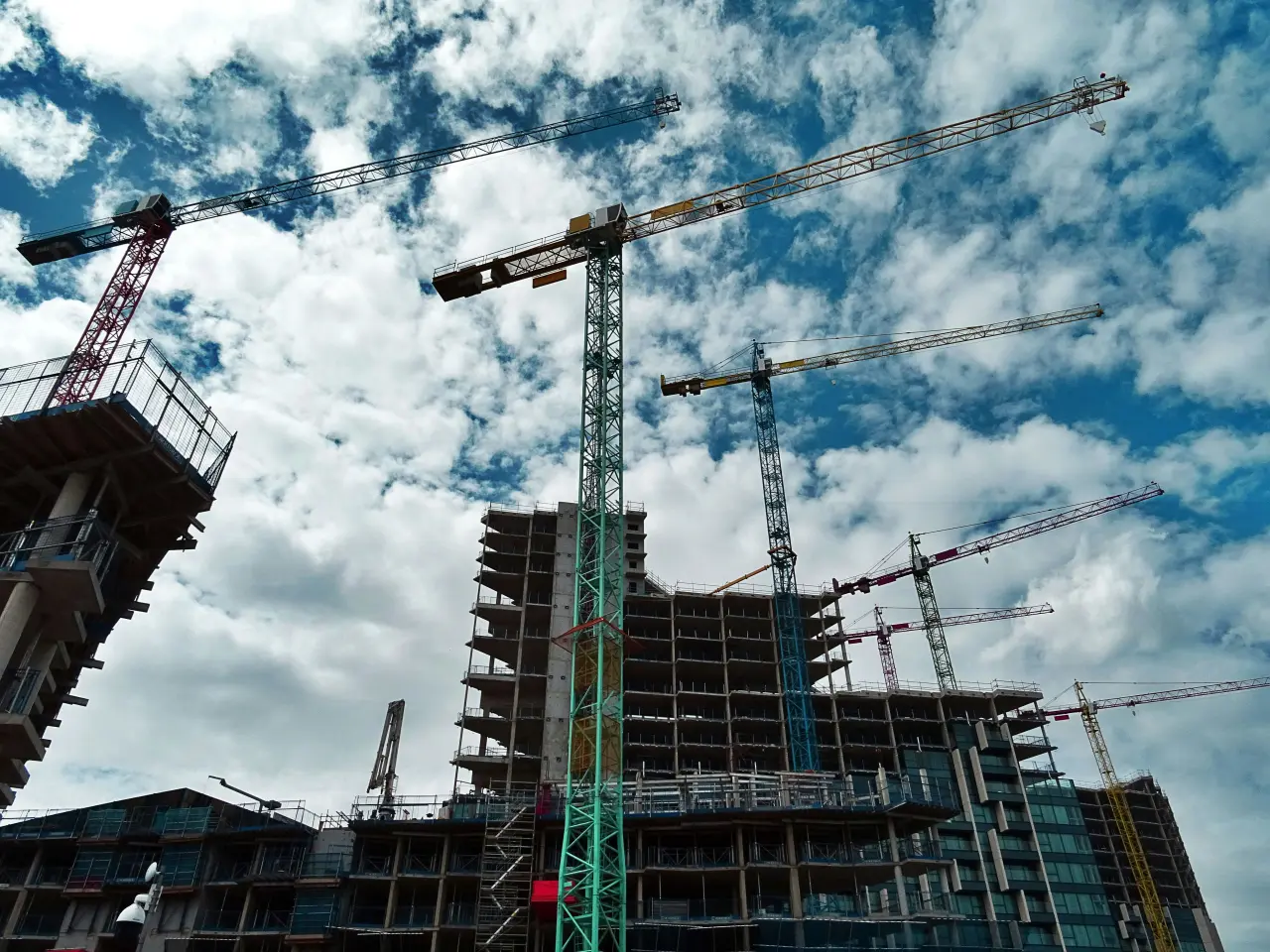 Multiple tower cranes operating around a high-rise building under construction, set against a partly cloudy sky.