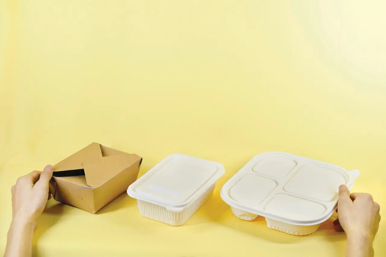Three takeaway food containers on a yellow background, with hands holding the left brown cardboard box and the right white plastic container.